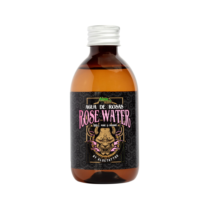 Un flacon en verre brun avec un bouchon argenté étiqueté "Eau de rose Aloe Tattoo 250 ml" comporte une étiquette noire ornée d'une illustration de rose et d'un texte décoratif rose et blanc.