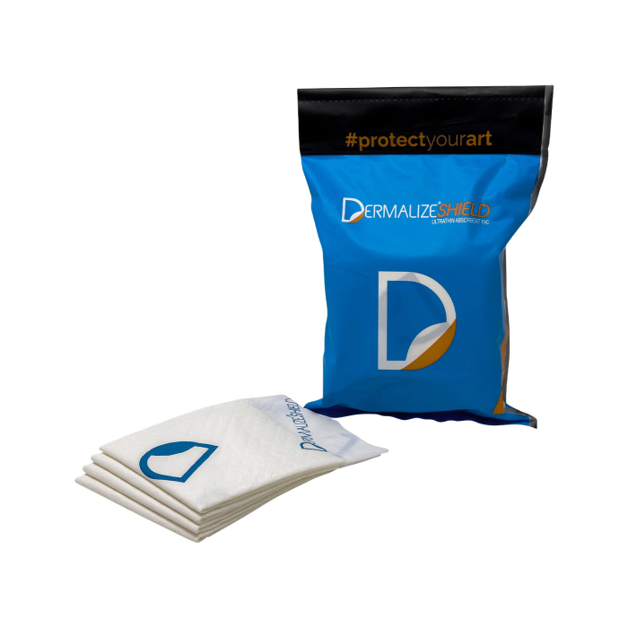 Un Dermalize SHIELD - Compresses absorbantes ultrafines - Boîte de 10 se tient debout, avec l’inscription « #protectyourart », à côté de quatre tampons blancs pliés portant un logo bleu « D », disposés devant l’emballage.