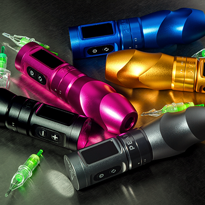 Una collezione di cinque macchine per tatuaggi rotative nei colori nero, grigio, rosa, arancione e blu, disposte su un tavolo con cartucce per tatuaggi accanto.